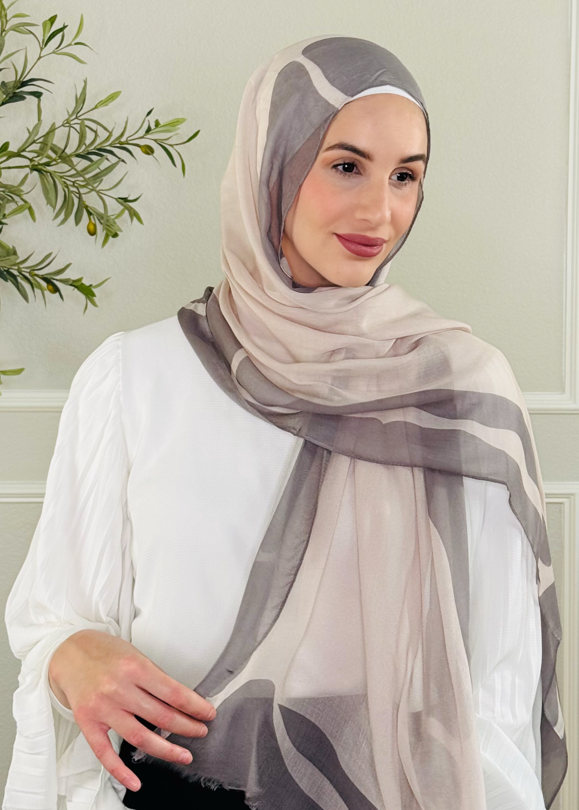 Woman wearing a gray and beige hijab in a room with a plant and white wall.