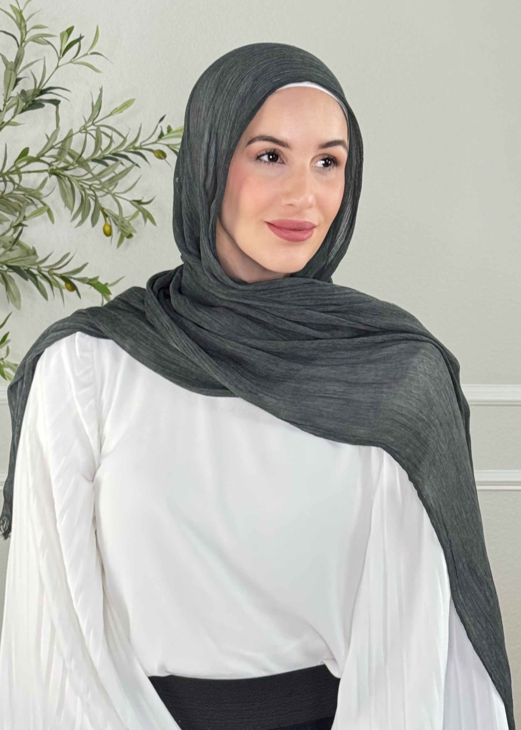 Woman wearing a dark gray hijab and white blouse against a light gray wall.